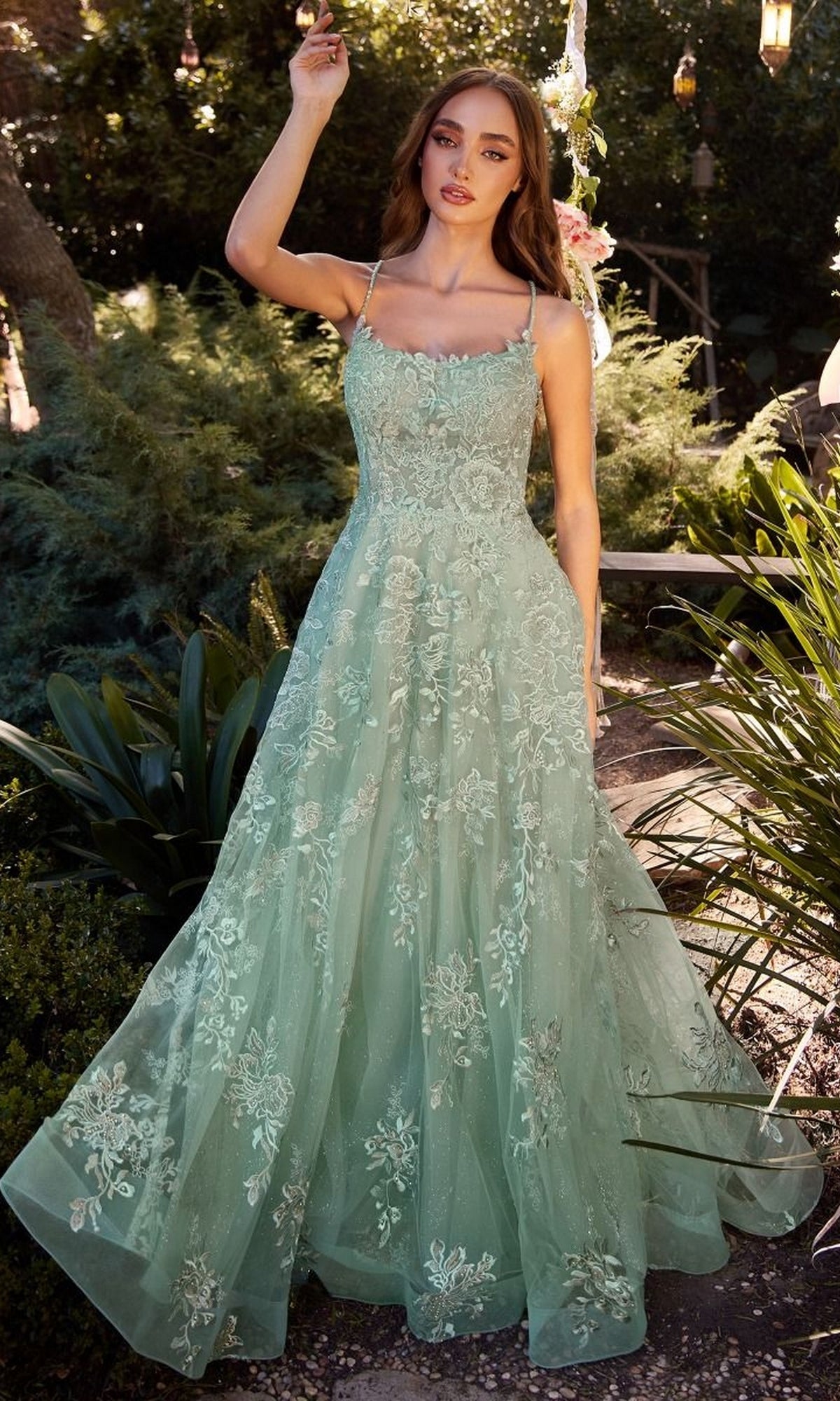 A woman stands outdoors in the Embroidered Prom Ball Gown: Andrea & Leo A1248, a light green, floral embroidered lace dress with spaghetti straps, surrounded by greenery and sunlight.